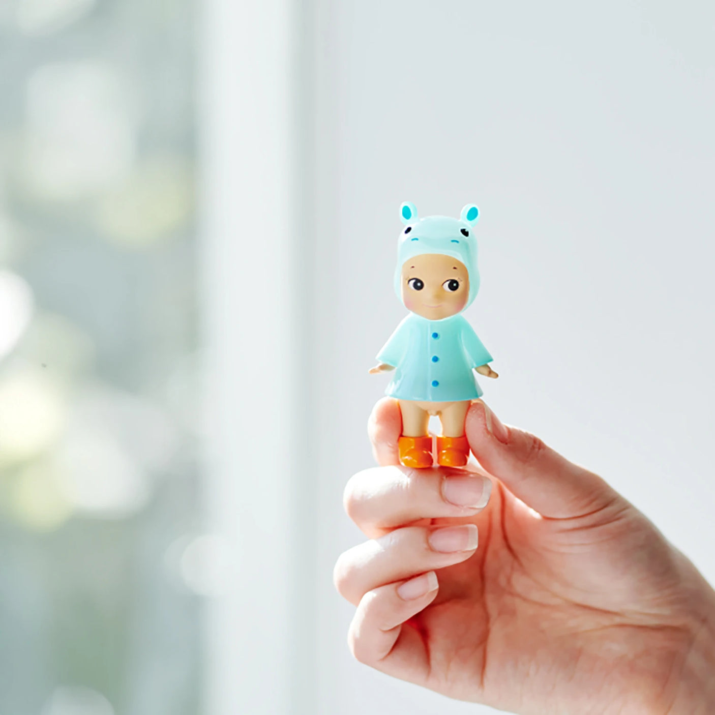 A hand holds a Sonny Angel Rainy Day Blind Box - Limited Edition doll by Sonny Angel, dressed in a light blue animal-themed hooded outfit and orange boots, set against a softly blurred background.