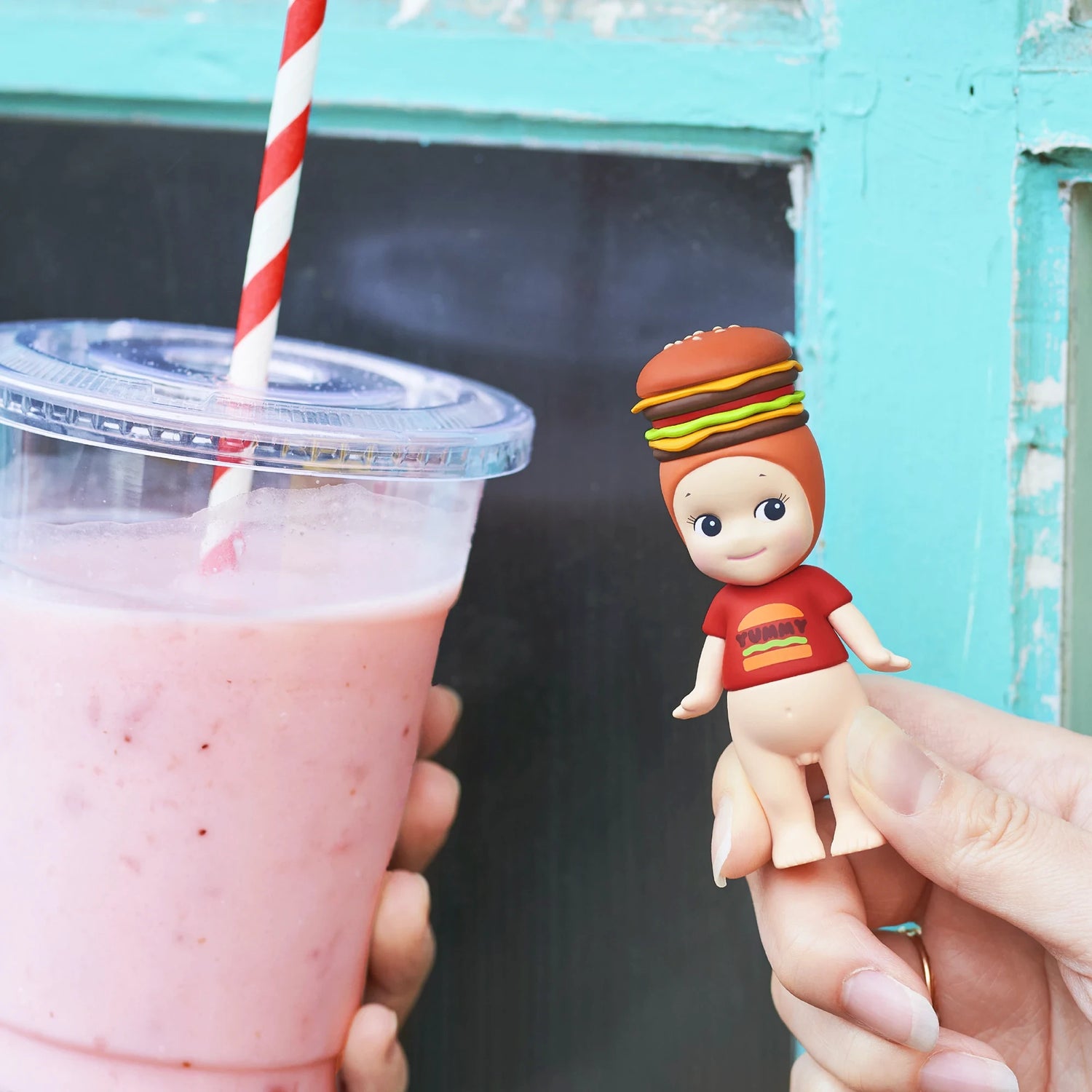 A hand holds a pink smoothie with a striped straw beside a Sonny Angel Snack Series - Blind Box mini-figure by Sonny Angel, wearing a hamburger hat and red shirt, set against a blue-green window frame.
