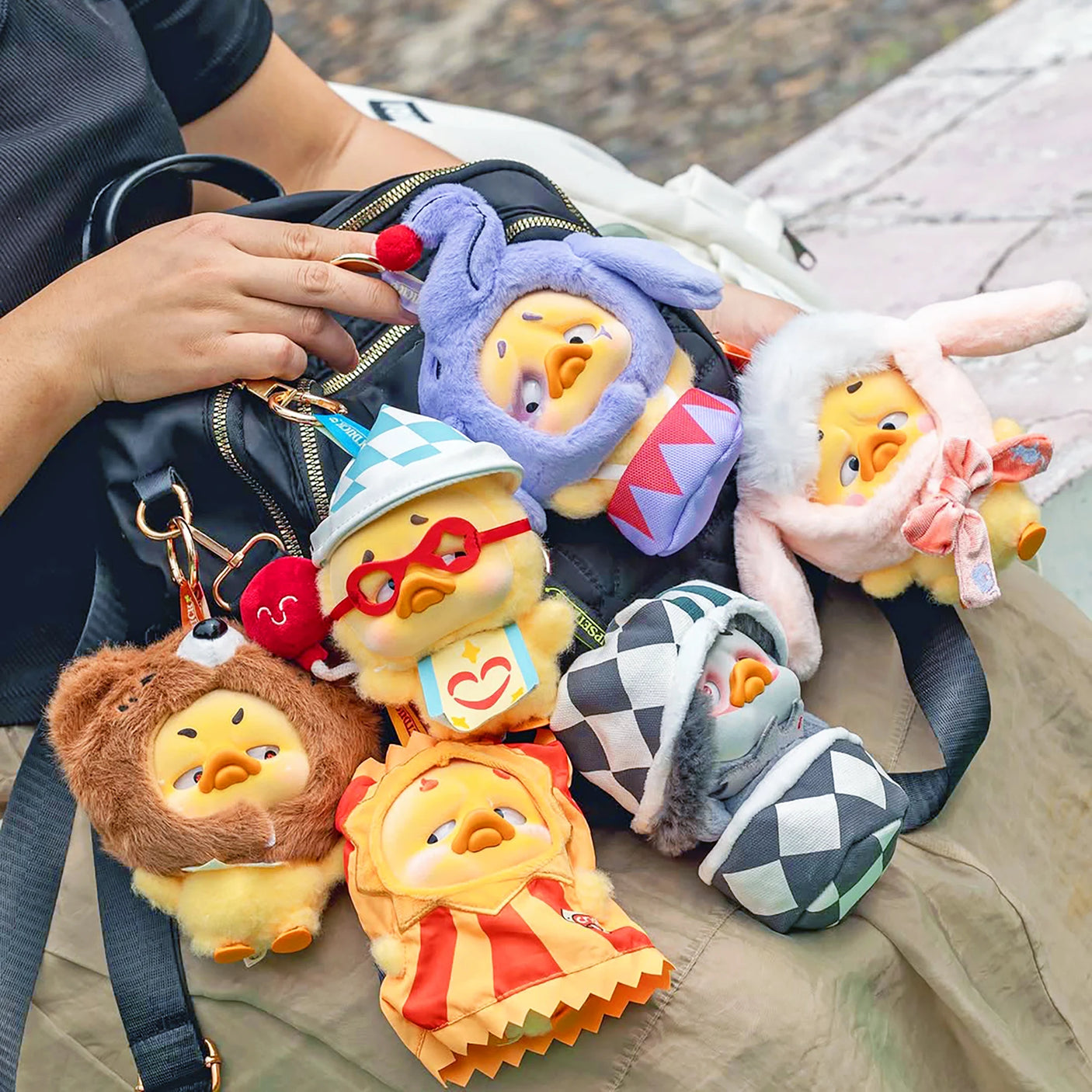 Vinyl plush duck keychains in mixed outfits, clipped to a bag strap.