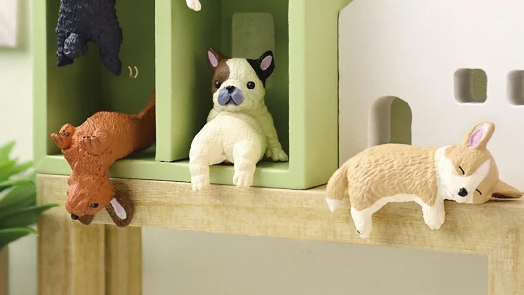 Three miniature dog figurines rest on and near a wooden shelf, with two inside a green structure and one lying on the edge.