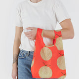 A person holds a vibrant Baby Baggu Reusable Bag - Yubari by Baggu, featuring a playful cantaloupe pattern, perfectly complementing their white t-shirt and blue jeans.