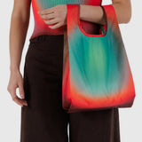 A person carries a Baby Baggu Bag x Julia Heuer - Rain, made from recycled nylon, featuring a colorful gradient. They're dressed in a matching Julia Heuer top and dark pants for an effortlessly stylish look.