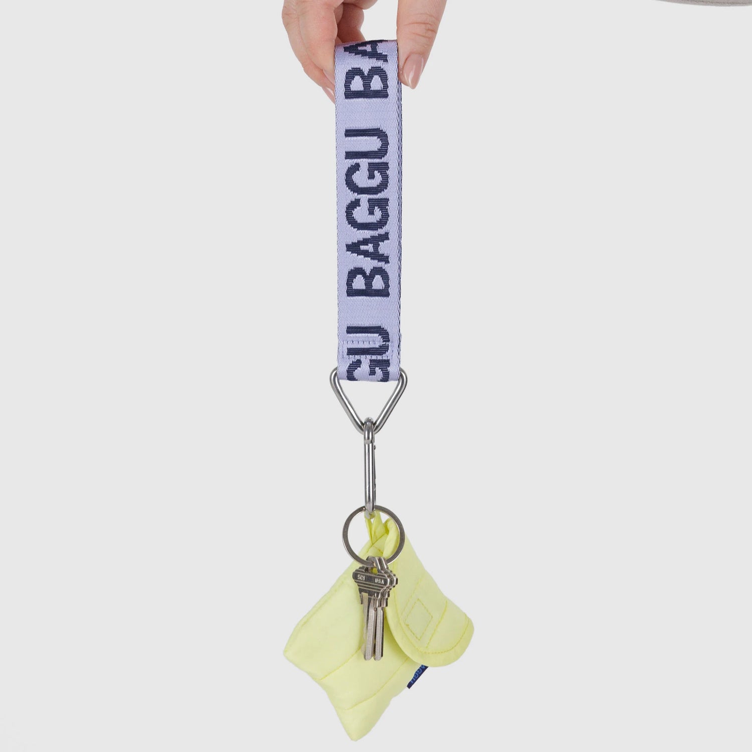 A hand holds the Baggu Logo Keychain - Dusty Lilac with two keys and a small pouch, attached to a white strap labeled "BAGGU" in blue letters.