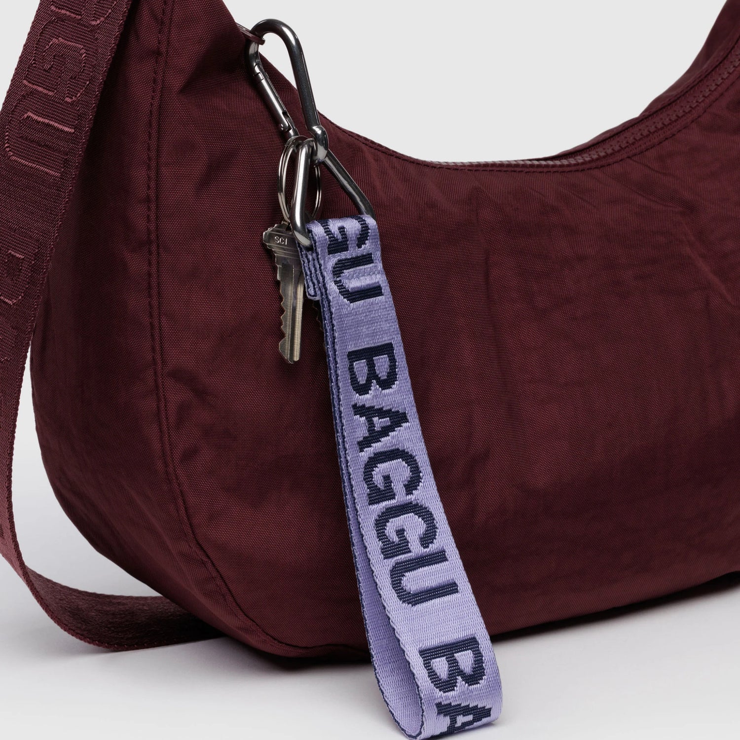 A silver key and a Baggu Logo Keychain in Dusty Lilac dangle from the zipper pull of a maroon bag.