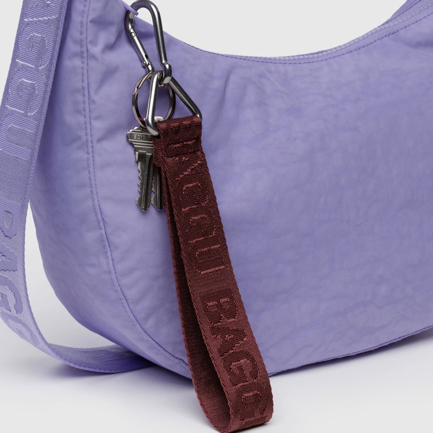 A close-up of a lavender shoulder bag with a purple strap, featuring the Baggu Logo Keychain in Mahogany by Baggu and a set of metal keys attached to the bag.