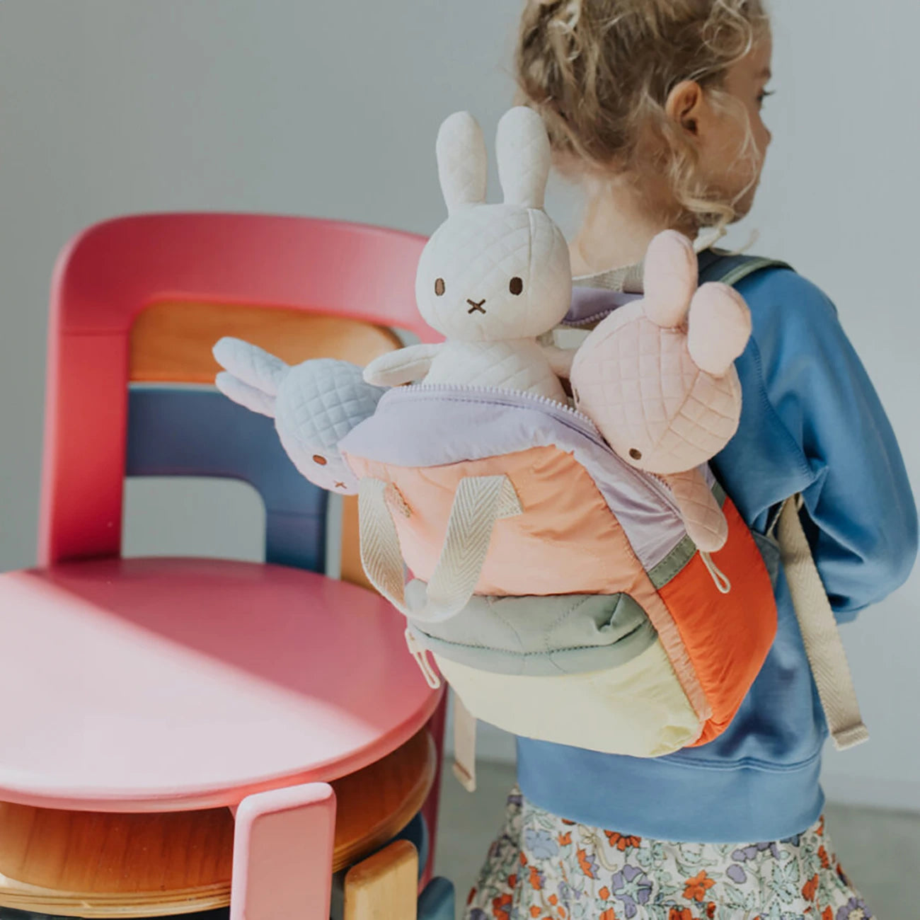 A child with a vibrant, handcrafted backpack filled with Miffy Quilted Plush Gift Box toys stands beside a pink and wooden chair.