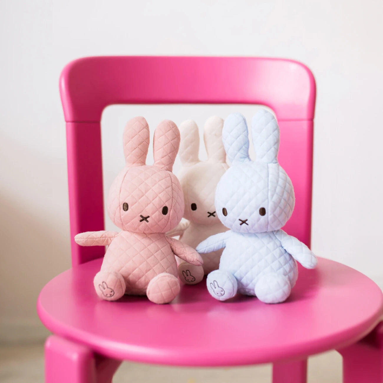 A vibrant pink chair holds a trio of eco-friendly Miffy quilted bunny plush toys, each sitting at 23cm tall, in colors of pink, white, and ice blue.