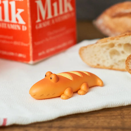 A small orange alligator figurine from Dreams Inc's Animal Bakery Blind Box - Vol 2, shaped like a loaf of bread, sits on a white cloth next to baguette slices and a milk carton.