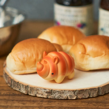 A small, orange rhino pastry figurine from Dreams Inc’s Animal Bakery Blind Box Vol 2 sits in front of three bread rolls on a wooden surface—ideal for animal-themed bakery or treat displays.