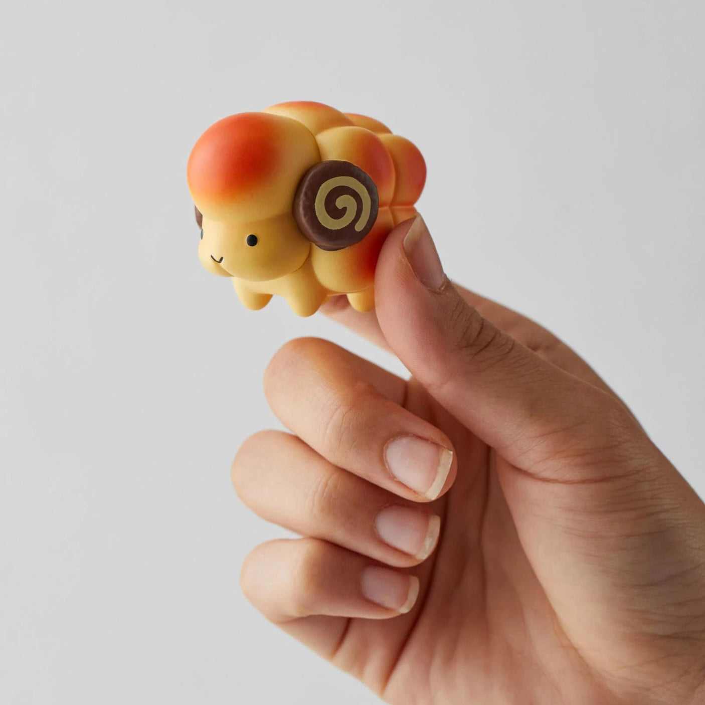 A hand holds a small toy sheep with a round body, orange and yellow coloring, and spiral ear patterns. This delightful surprise from the Animal Bakery Mini Figure Blind Box series by Dreams Inc is part of their collectible range.