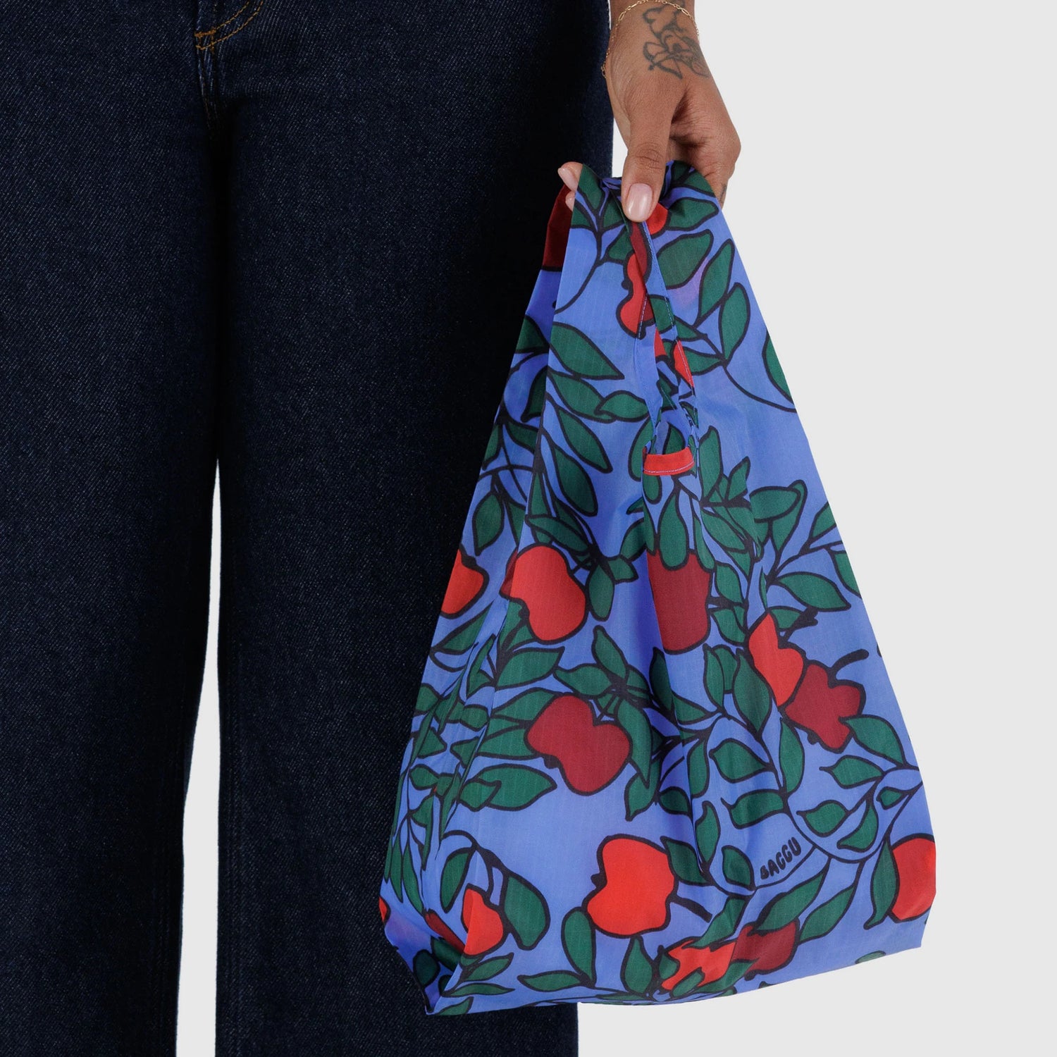 A person holding the Baggu Baby Baggu Bag - Apple Tree, a reusable shopping bag featuring a red and green apple tree print on a blue background.