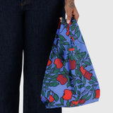 A person holding the Baggu Baby Baggu Bag - Apple Tree, a reusable shopping bag featuring a red and green apple tree print on a blue background.