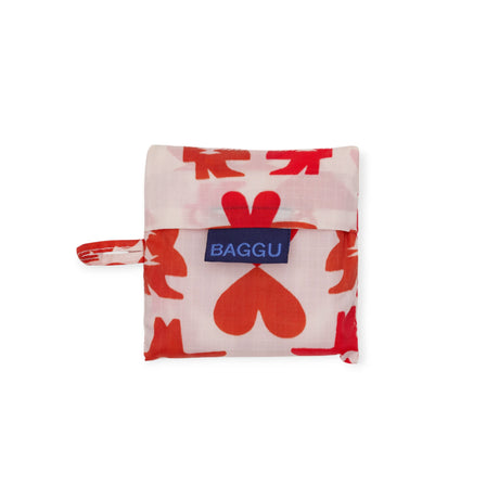 The folded Baby Baggu Bag - Cowgirl by Baggu features a red and pink heart and handprints pattern, secured with a flap and label—a cute, reusable accessory for any cowgirl bag collection.