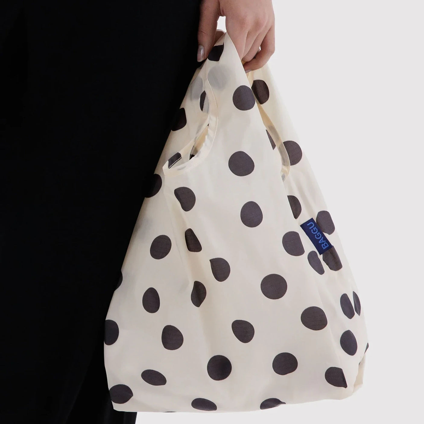 Cream baby tote bag with black polka dots, held by hand with blue logo patch