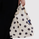 Cream baby tote bag with black polka dots, held by hand with blue logo patch