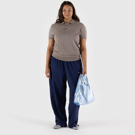 A person wearing a beige polo shirt and blue pants holds a Baggu Baby Baggu Bag in Metallic Light Blue, standing against a plain white background.