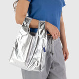 A person in a blue shirt and gray pants is holding a Baby Baggu Bag - Metallic Silver by Baggu against a plain background.