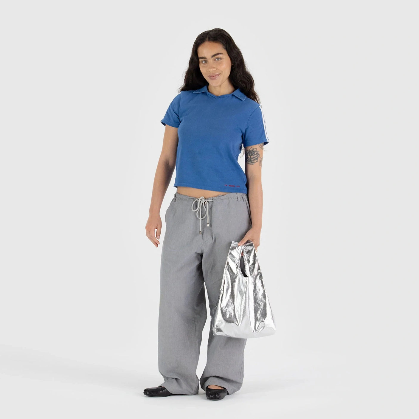 An individual with long hair in a blue shirt and gray pants stands against a simple background, holding a Baggu Baby Baggu Bag in Metallic Silver.