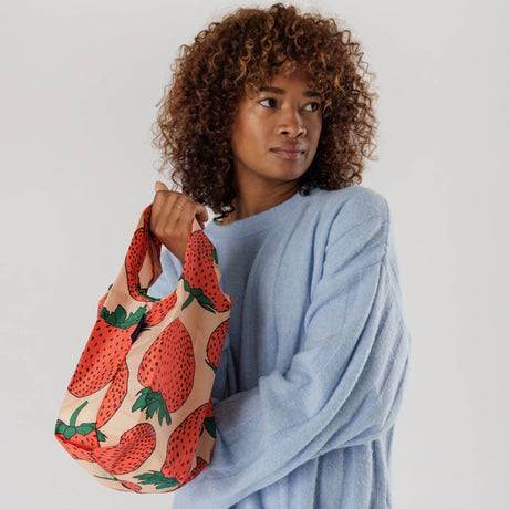 A person wearing a blue sweater holds a Baggu Baby Baggu Reusable Bag in the strawberry pattern against a plain background.