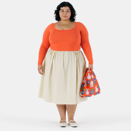 A person in an orange long-sleeve top and cream skirt holds a red Baby Baggu x Miffy Flower reusable bag by Baggu featuring a cartoon bunny print, standing against a plain white background.