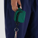 A hand holds the Baggu Fanny Pack Charm - Cypress, crafted from recycled nylon and attached to a keychain with keys. Its vibrant green contrasts beautifully against navy blue pants, adding a stylish touch for those on the go.