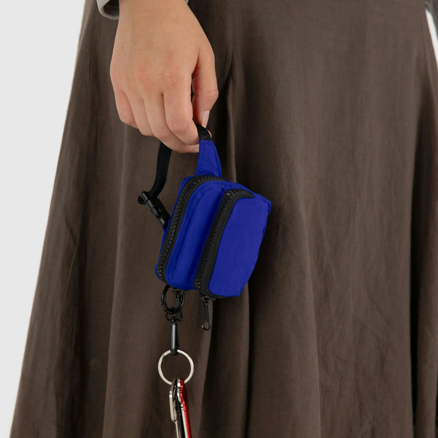 A person holds a Baggu Fanny Pack Charm featuring a small blue pouch, paired with a brown skirt. The recycled heavyweight nylon adds both style and sustainability to the outfit.