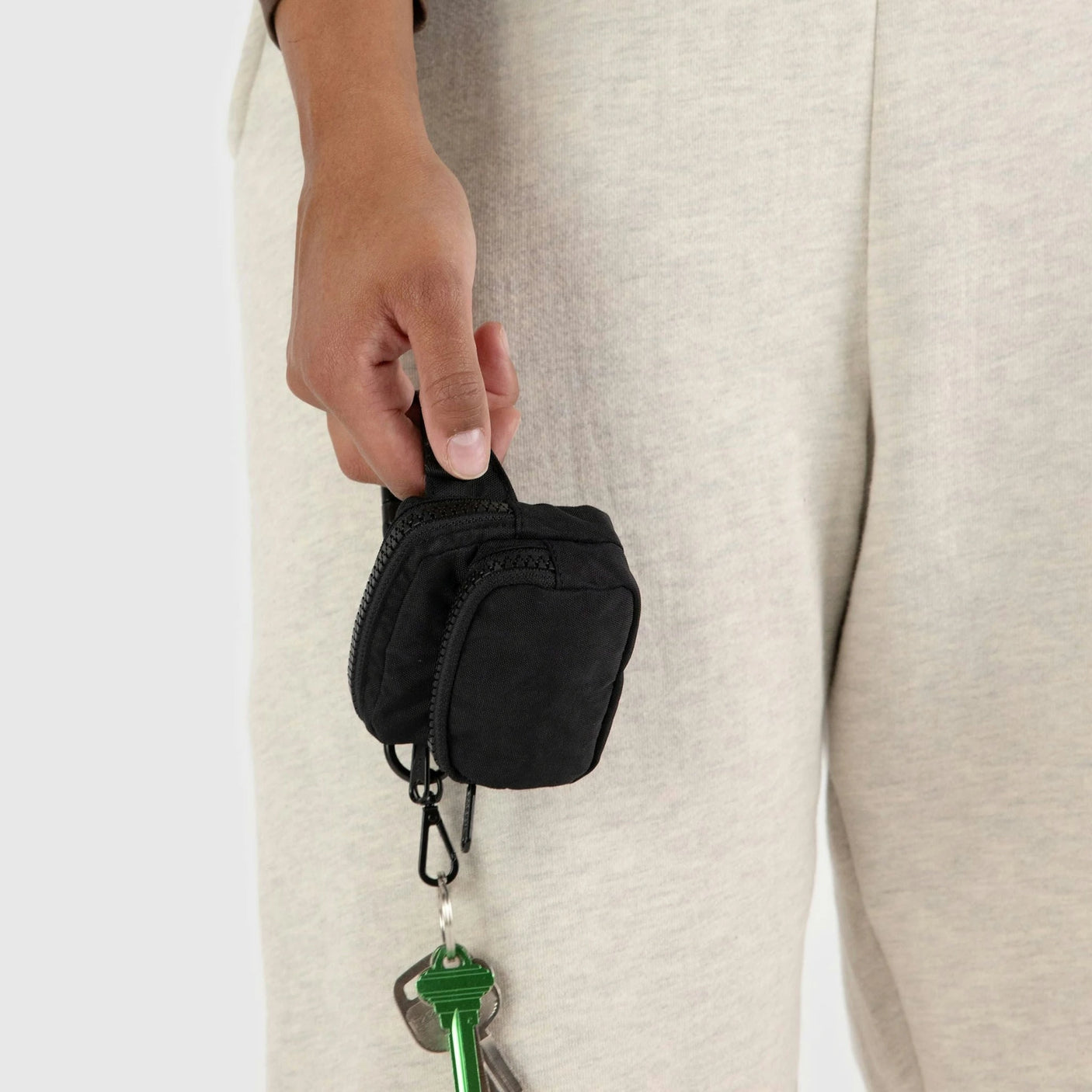 A hand holds a small black pouch with a zipper, similar to a Baggu Fanny Pack Charm, attached to keys against light-colored pants.