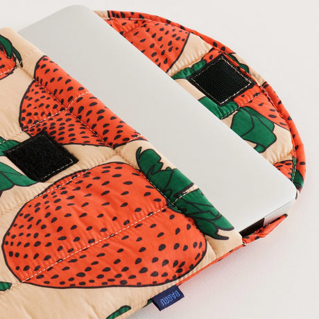 A silver laptop partially slides into the Baggu Puffy Laptop Sleeve 13"/14", which is crafted from recycled materials and features a bold red strawberry print with black velcro closures.