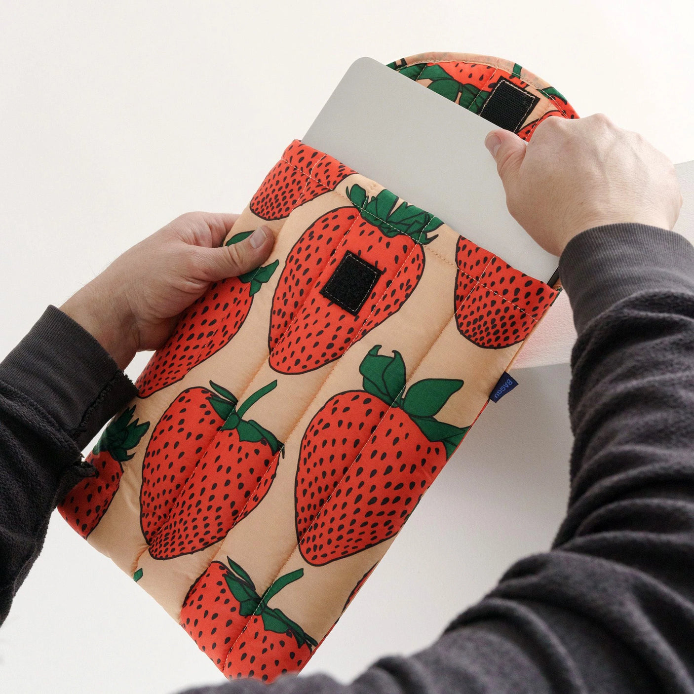 A person is carefully sliding their tablet into the Baggu Puffy Laptop Sleeve 13"/14" - Strawberry, a charming sleeve thoughtfully crafted from recycled materials.