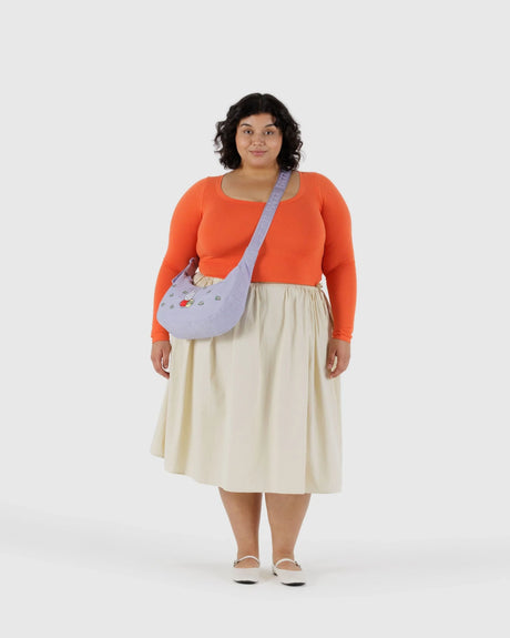 A woman faces forward, wearing an orange long-sleeve top, cream skirt, white shoes, and the Baggu Miffy x Baggu Medium Nylon Crescent crossbody bag in lavender with small embroidered details.