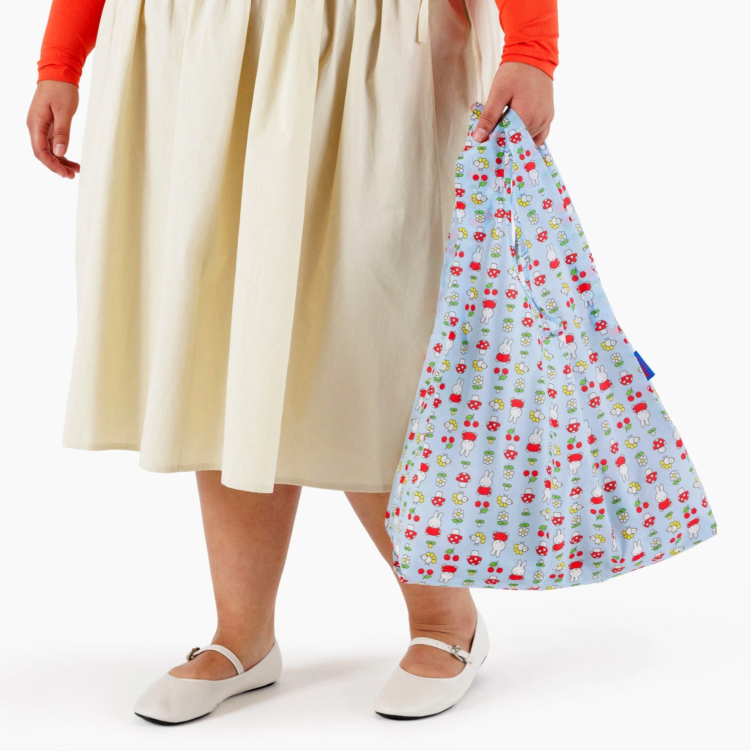 A person in a beige skirt and white shoes holds a Baggu Standard Baggu x Miffy Garden reusable bag with a colorful mushroom pattern.