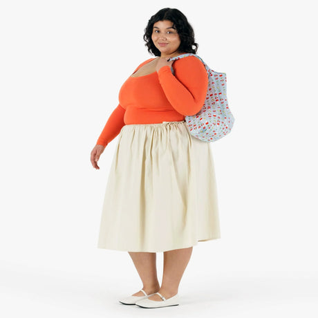 A woman with curly dark hair wears an orange long-sleeve top, cream skirt, white shoes, and carries the Baggu Standard Baggu x Miffy Garden reusable bag over her shoulder.