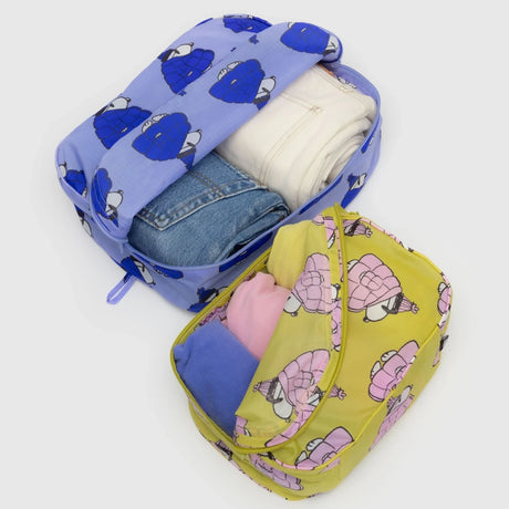 The Peanuts x Baggu - Packing Cube Set by Baggu includes two recycled nylon zipper pouches: one blue with jeans and white pants, the other yellow with pink and purple clothes, both featuring delightful cartoon character prints.