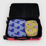 An open suitcase reveals two packing cubes from the Peanuts x Baggu Packing Cube Set: a blue one with a shell pattern and a yellow one. Made from recycled nylon, these cubes add a playful touch to your travels.