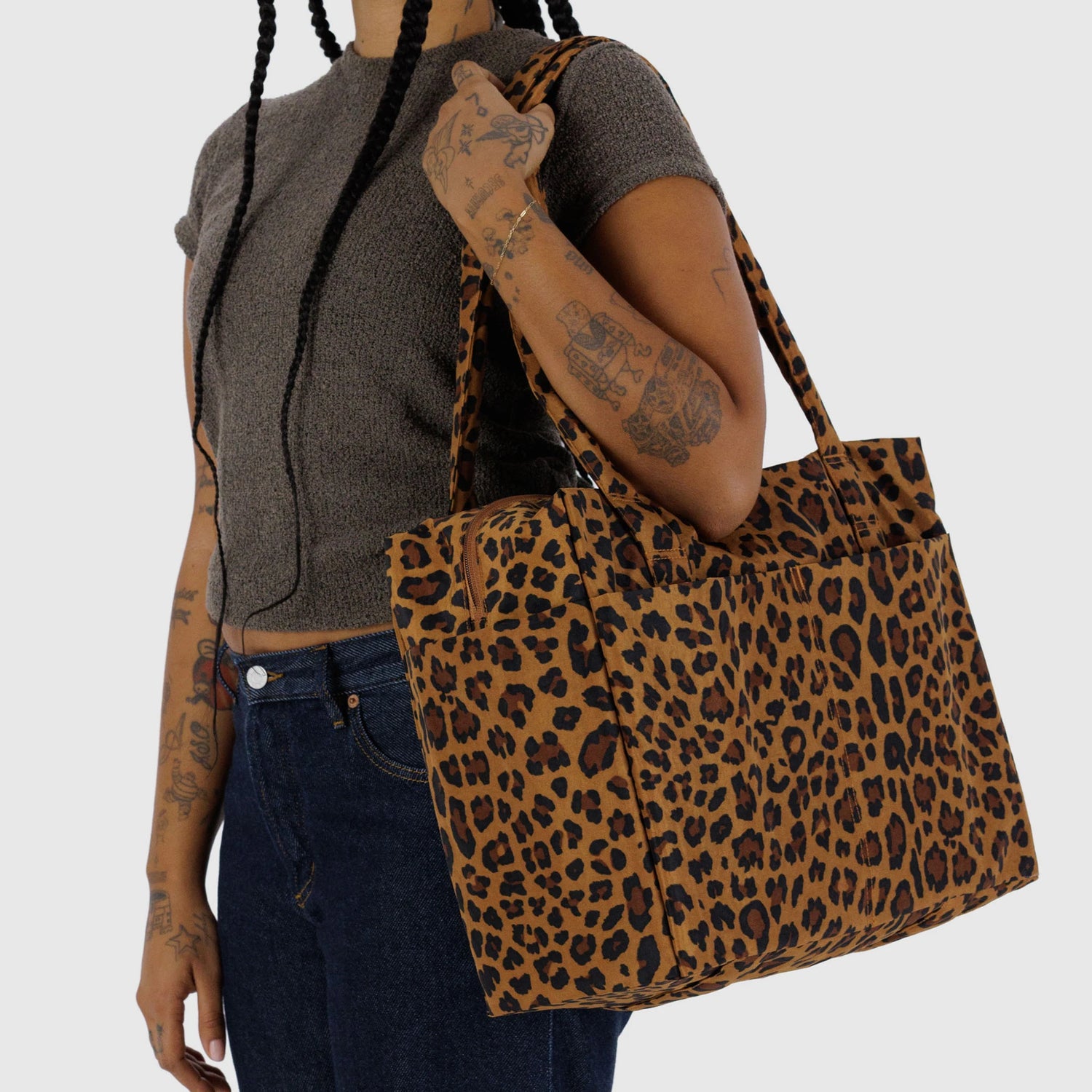 A person with braids and tattoos holds the Baggu Small Cloud Carry-on - Leopard by Baggu, styled with a short-sleeve brown top and dark jeans.