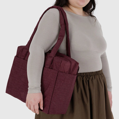 A person wearing a grey long-sleeve top and brown skirt carries the Baggu Small Cloud Carry-on in Mahogany by Baggu over their shoulder.
