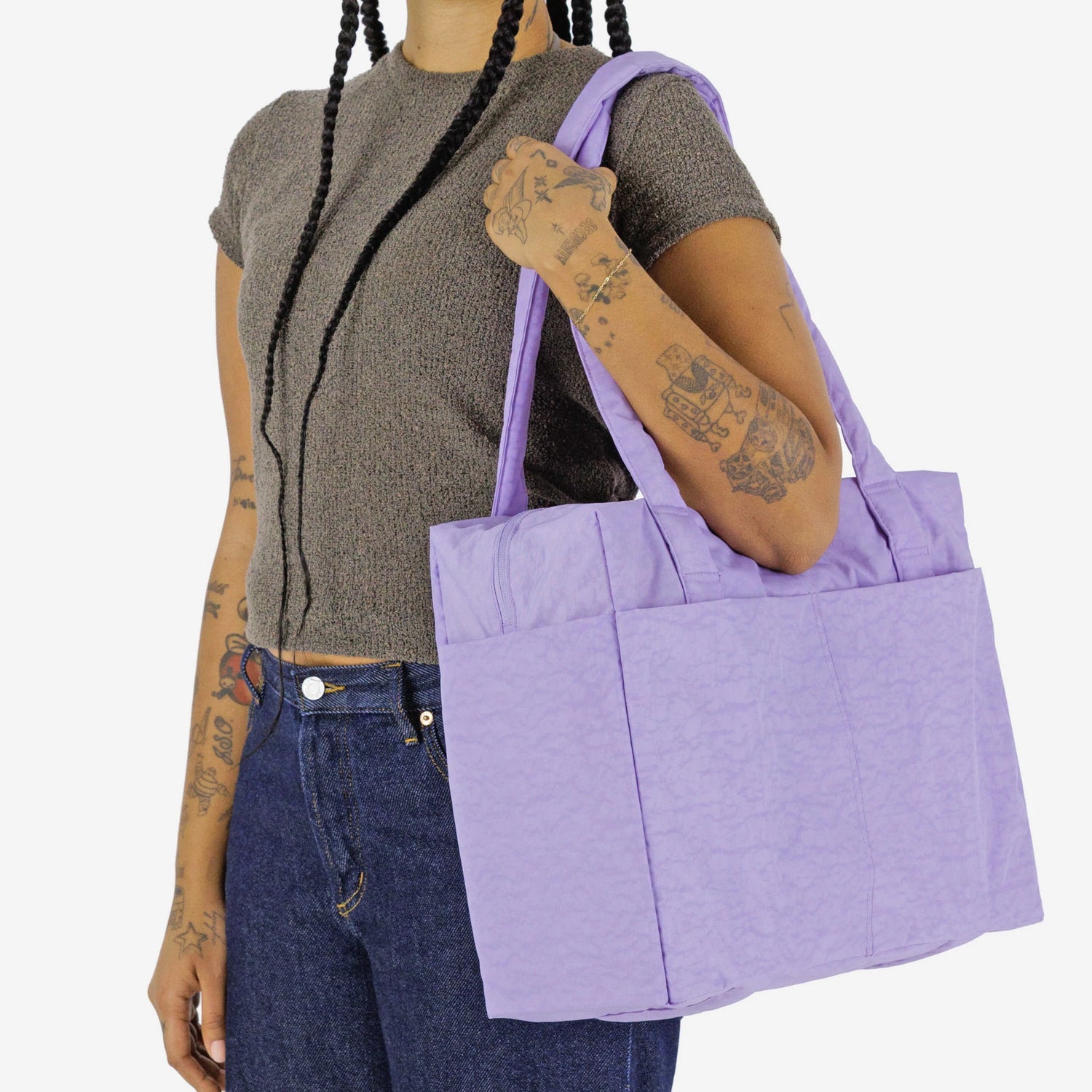 A person with braided hair and tattoos carries the Baggu Small Cloud Carry-on - Wisteria on their shoulder, styled with a short-sleeve knit top and blue jeans—an ideal underseat weekender from Baggu for chic, practical travel.