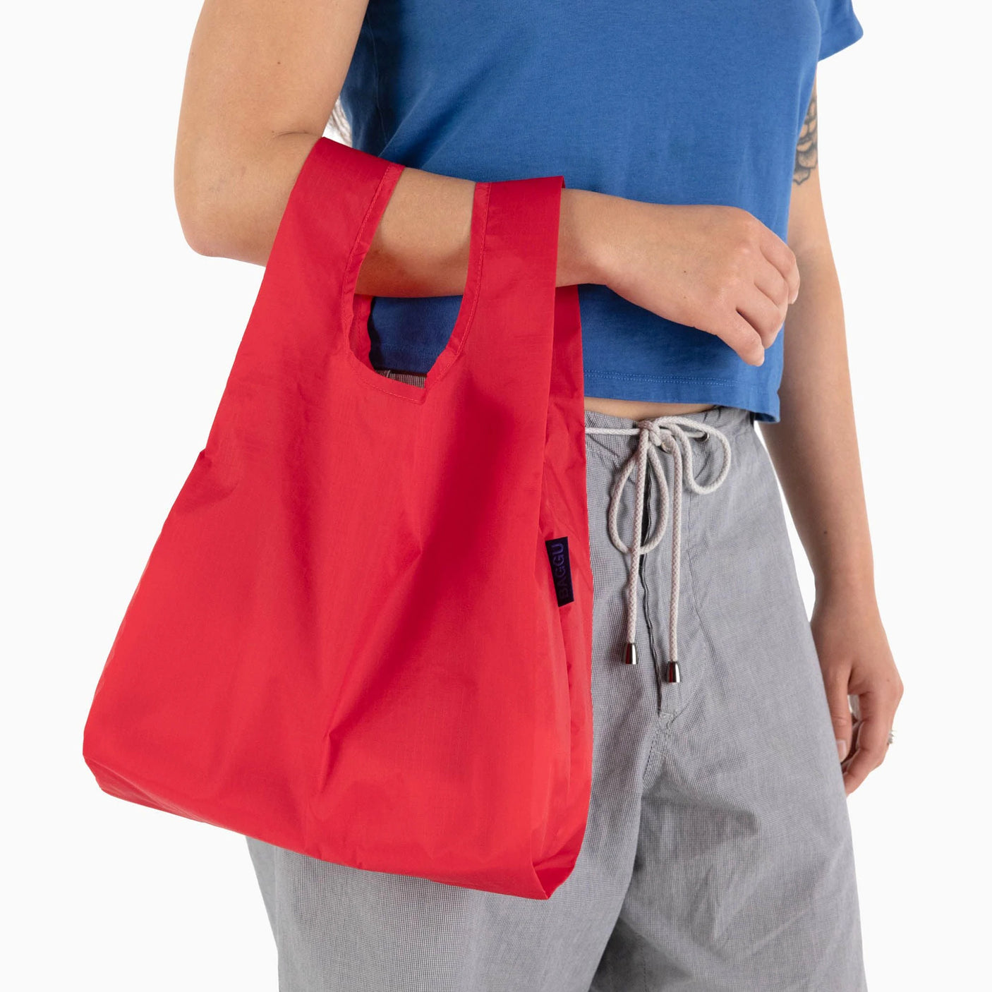 A person in a blue shirt and gray pants holds a Baggu Baby Baggu Reusable Bag in Candy Apple red, made from eco-friendly 100% recycled nylon.