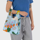 A tattooed individual is seen holding a vibrant Baby Baggu Reusable Bag in the "Table Cats" design, which showcases cats and floral patterns and is crafted from recycled nylon. They complement their look with a blue shirt and gray pants.