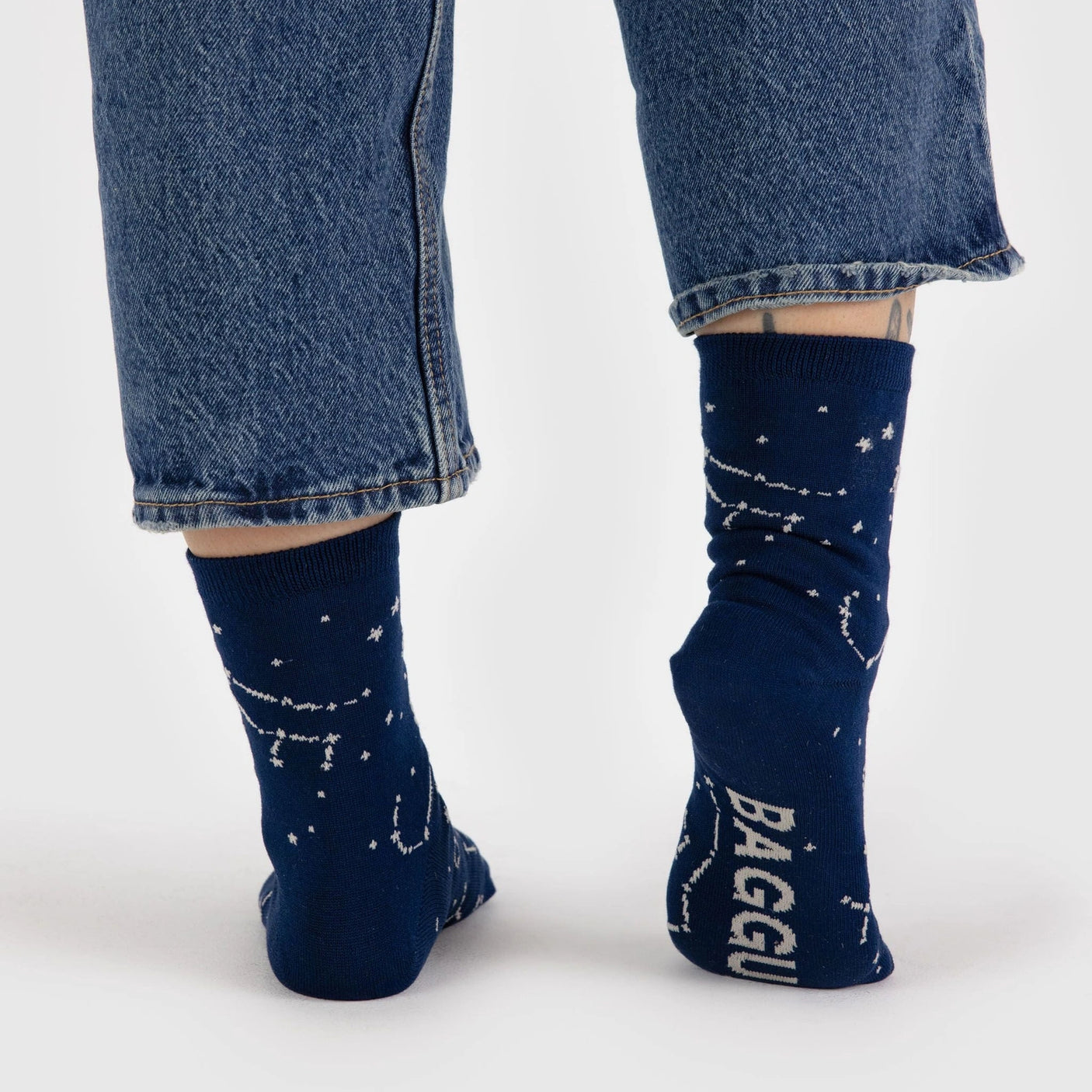 A person wearing the Baggu Crew Sock in the Constellation Midnight design, paired with rolled-up jeans, is shown from behind, highlighting the stellar pattern on their blue socks.