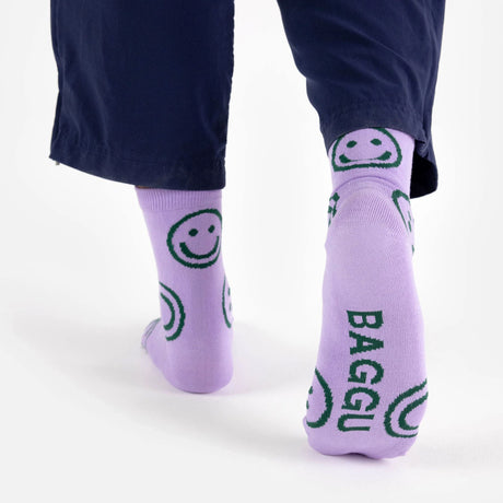 A person wearing Baggu Crew Sock - Lavender Happy, adorned with green smiley faces and the "BAGGU" text, complements their dark pants effortlessly. Made from soft bamboo rayon, these socks by Baggu bring a cheerful element to any ensemble.