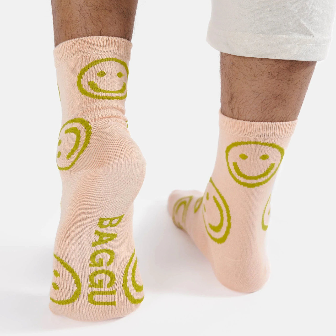 A person stands on a white surface, wearing Baggu Crew Sock - Light Pink Happy. These socks from Baggu are a true mood booster with their cheerful design featuring green smiley faces, brightening any day with comfort.