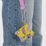 The Peanuts x Baggu Snoopy Puffer, a compact and stylish yellow pouch adorned with a pattern, features a purple strap with an attached carabiner clip, making it hang effortlessly from denim jeans and providing the perfect storage solution for your earbuds.