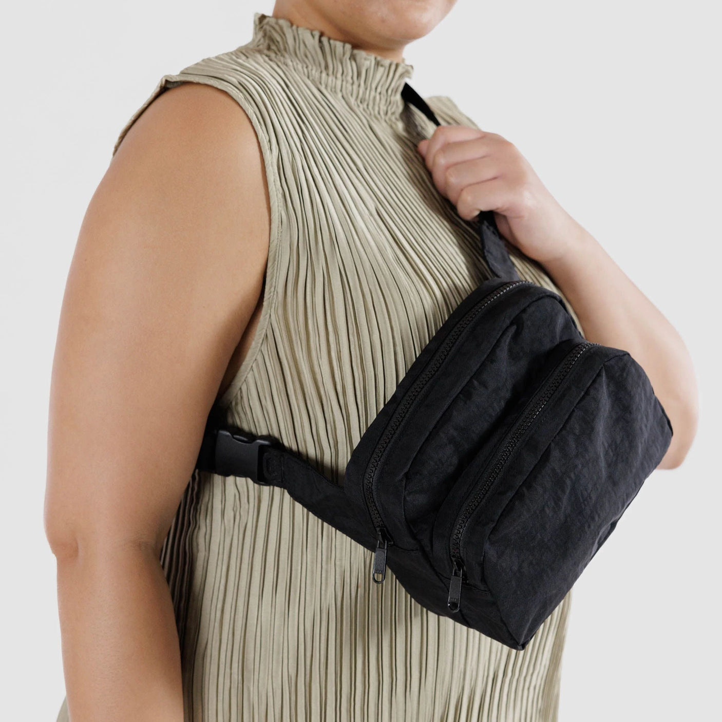 A person wearing a pleated top and the Baggu Fanny Pack - Black, a minimalist black fanny pack by Baggu made from recycled nylon with multiple zippered compartments, stands against a neutral background.