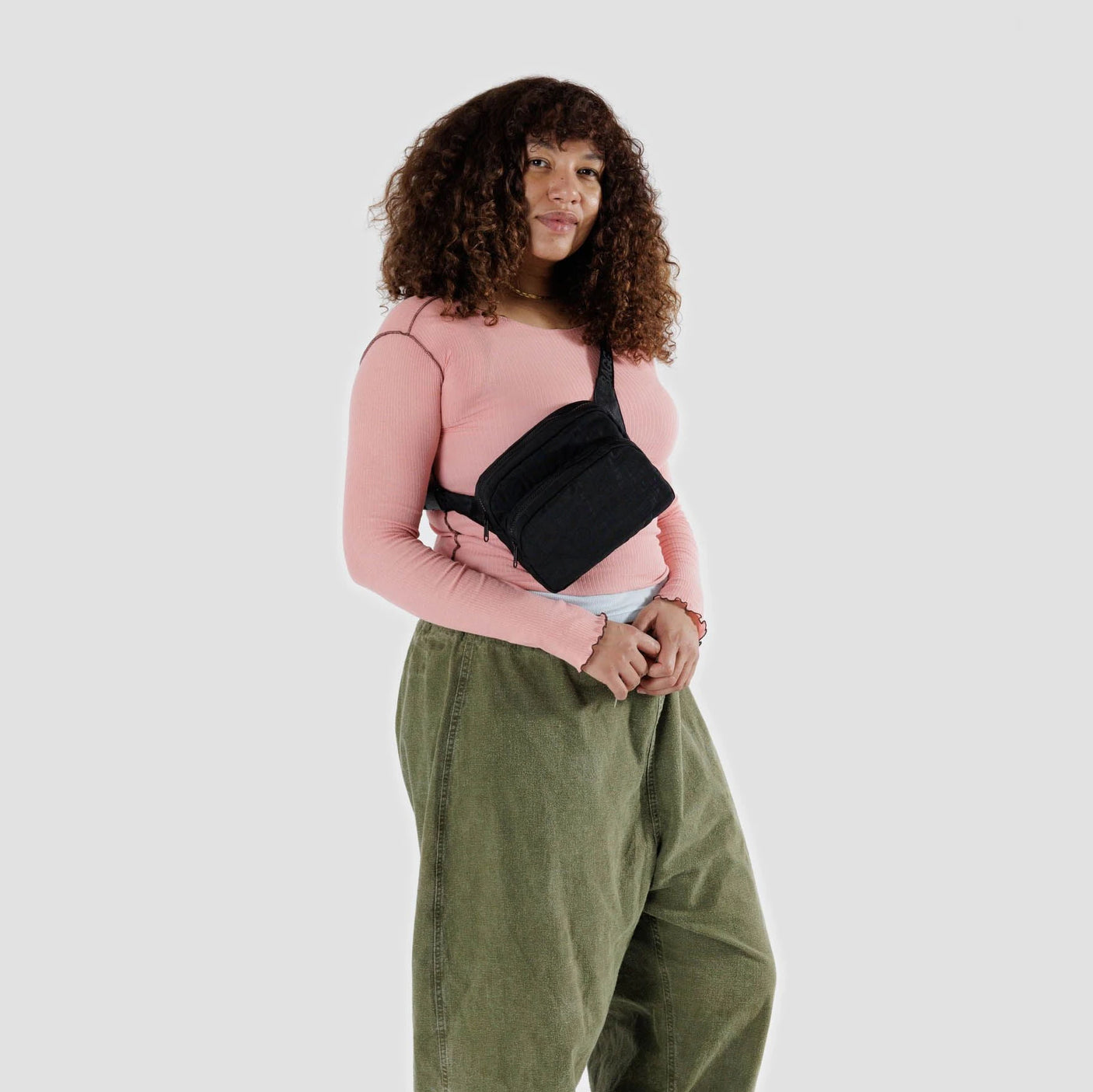 A person in a pink top and green pants stands against a plain background, sporting a sleek, minimalist black Baggu Fanny Pack crafted from recycled nylon.