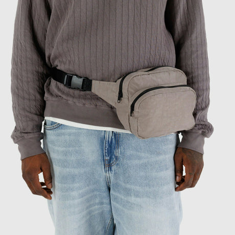 A person wearing a gray sweater and blue jeans completes their outfit with a Baggu Fanny Pack Dove, which features an adjustable strap made from durable nylon.