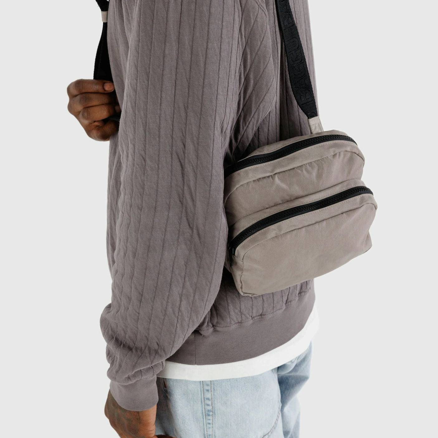 A person clad in a grey quilted jacket effortlessly holds the adjustable strap of the durable dove-colored Baggu Fanny Pack, with their jeans peeking through.