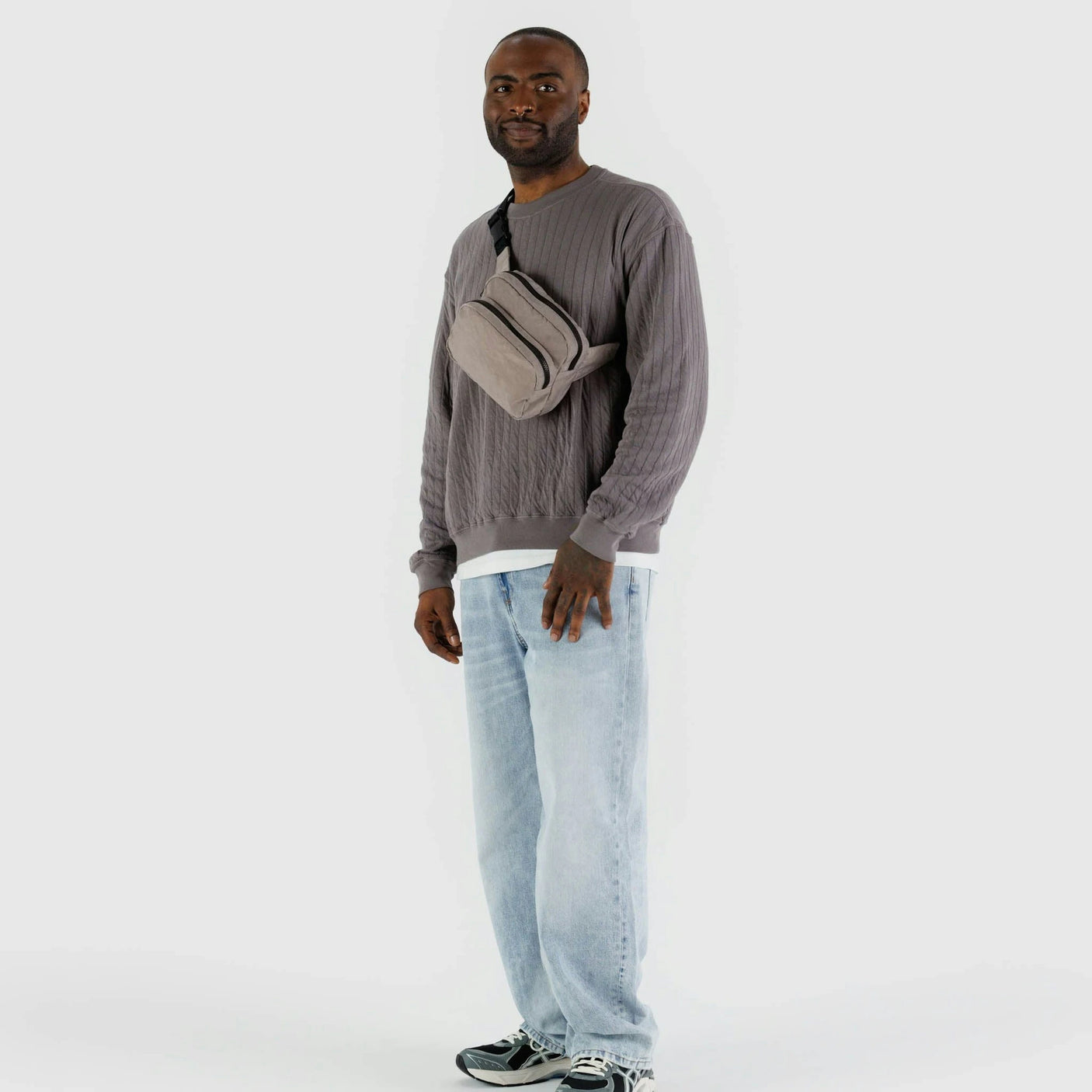 A person stands against a plain background wearing a gray sweater, light blue jeans, sneakers, and a Baggu Fanny Pack Dove with an adjustable strap.