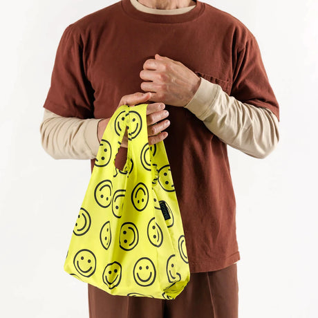 Person holding yellow Baby Baggu reusable bag decorated with large black smiling faces on white background