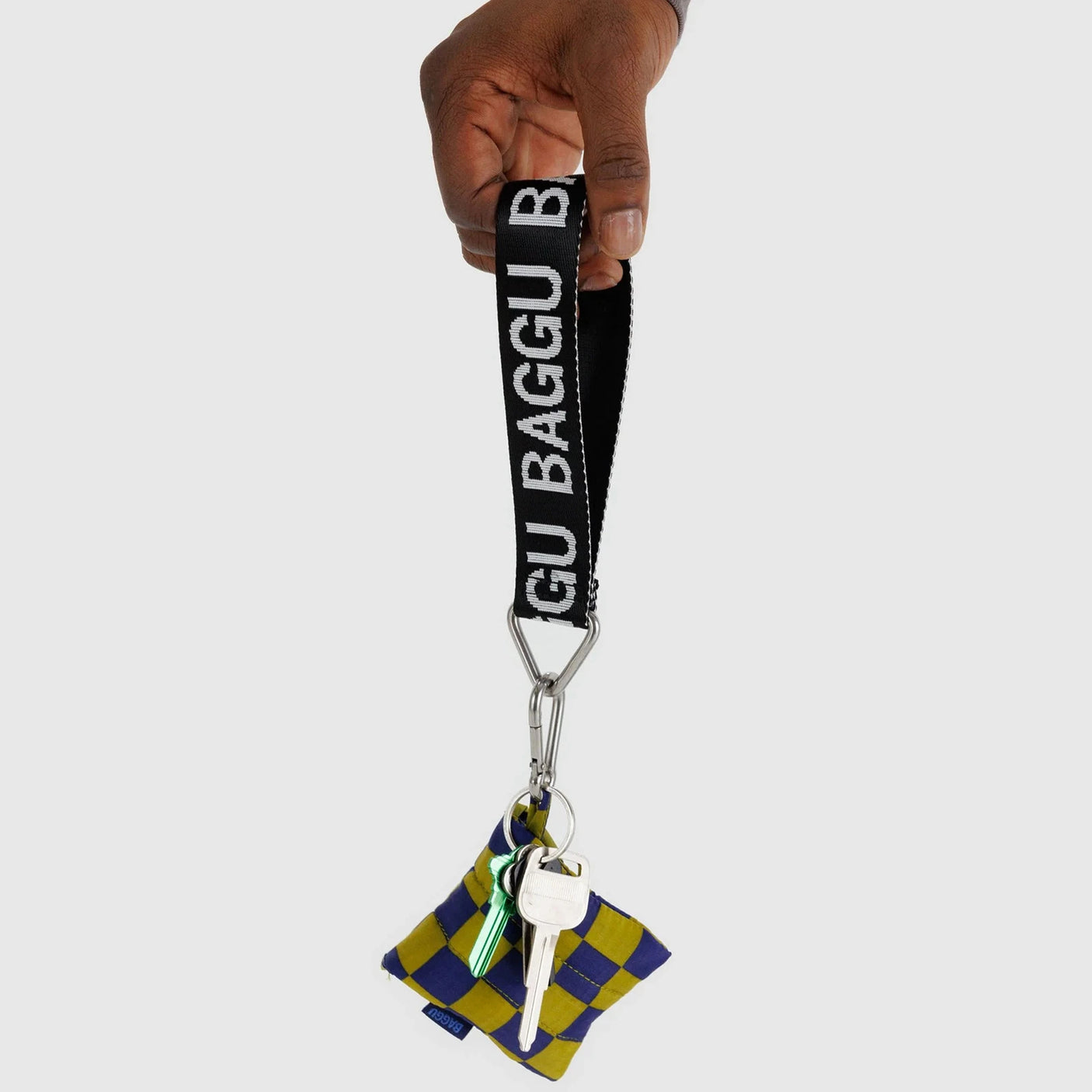 A hand holds the Baggu Keychain - Logo Strap, crafted from recycled nylon webbing, featuring the "BAGGU" brand on it. The strap is attached to a set of keys and a small, eco-friendly pouch with a blue and yellow checkered design.